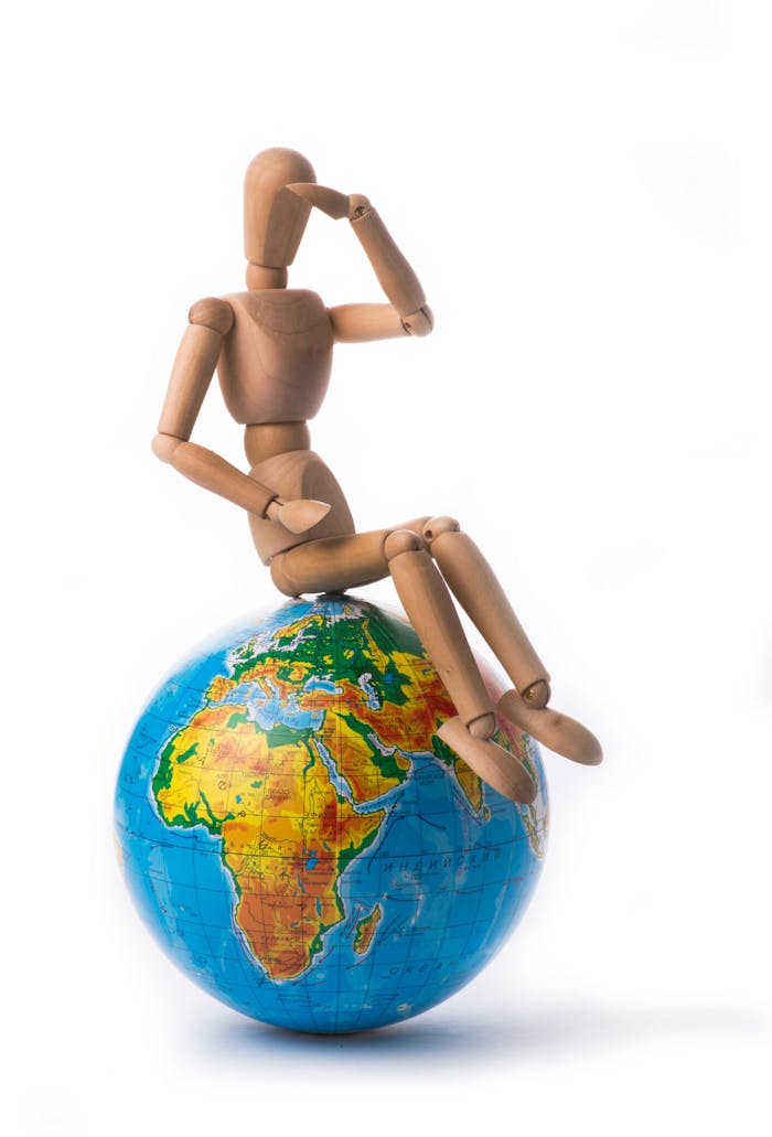 A wooden mannequin sits pondering on a colorful world globe against a white background.