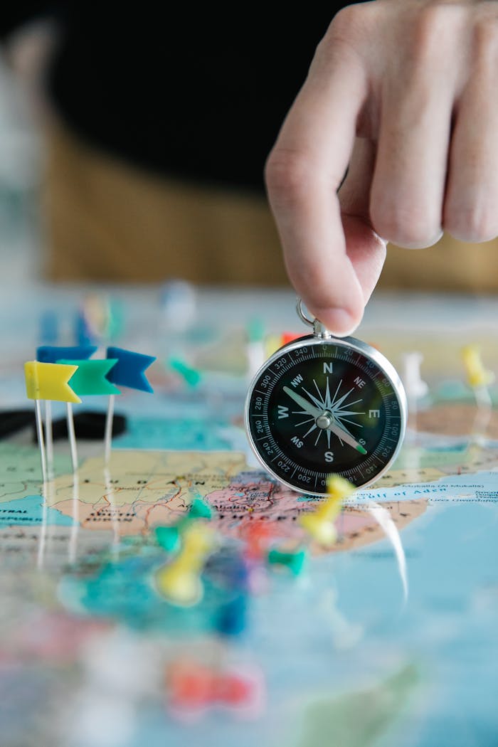 Close-up of hand holding compass over a map with colorful pins and flags, symbolizing travel planning.